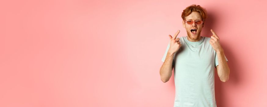 Young handsome man with red hair and beard pointing fingers at sunglasses, showing new glasses, standing over pink background.