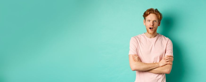 Intrigued redhead guy listening gossips, looking amazed and interested at camera, standing over turquoise background in casual t-shirt.