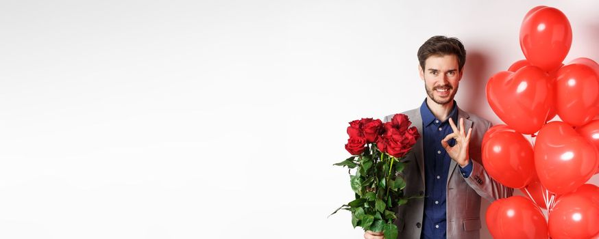 Young man in suit have all under control, showing OK sign and holding red roses, prepare romantic surprise with heart balloons and flowers on Valentines day, white background.