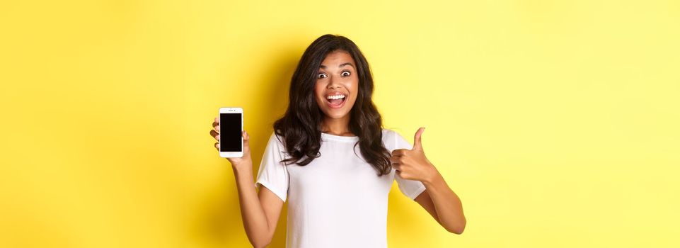 Portrait of excited african-american girl, showing smartphone screen and making thumbs-up, recommending something cool, standing over yellow background.