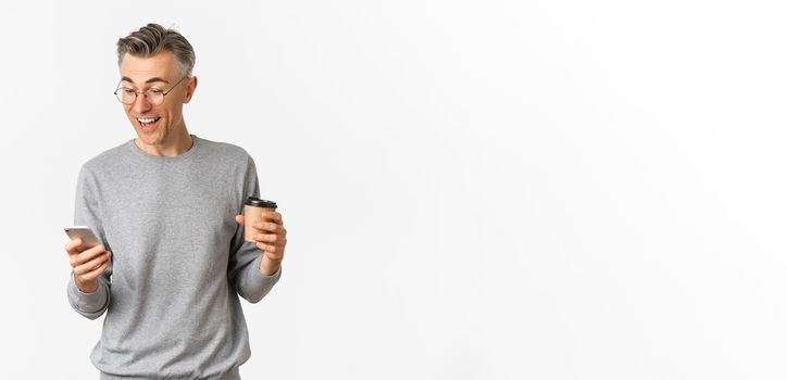 Image of handsome middle-aged man in grey sweater and glasses, drinking coffee and looking happy at smartphone screen, reading something online, standing over white background.