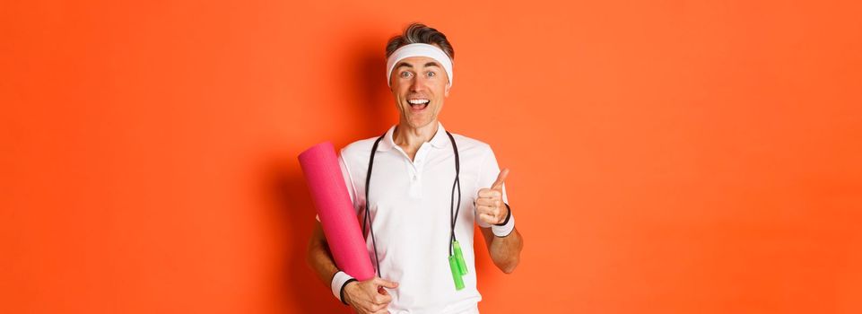 Concept of workout, gym and lifestyle. Cheerful middle-aged fitness guy, holding skipping rope and yoga mat, showing thumbs-up and smiling satisfied, standing over orange background.