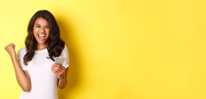 Image of sassy african-american girl in white t-shirt, making fist pump gesture and holding smartphone, celebrating achievement, standing over yellow background.