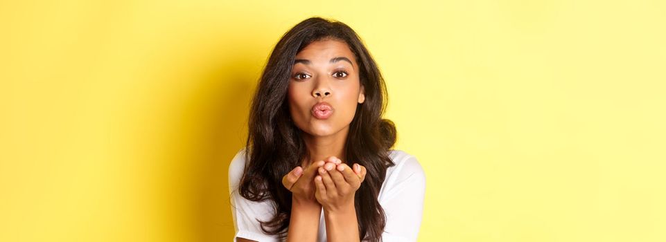 Close-up of beautiful and romantic african-american woman, pucker lips and sending air kiss, standing against yellow background.