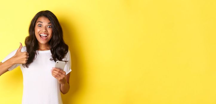 Portrait of amazed african-american girl, showing thumbs-up and using mobile phone, praising cool app, standing over yellow background.