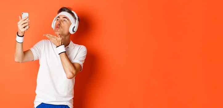 Portrait of attractive middle-aged man in gym uniform, wearing headphones, sending air kiss at mobile phone camera, taking selfie or having video call, standing over orange background.