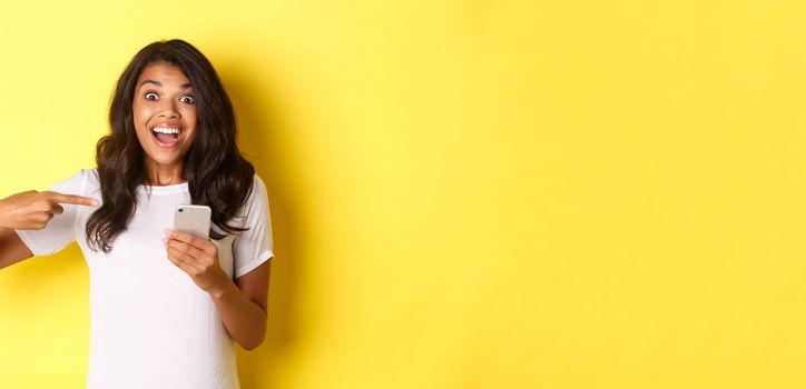Portrait of beautiful african-american girl, pointing finger at mobile phone to show something exciting, smiling amazed and looking at camera, standing over yellow background.