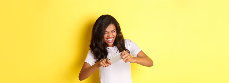Portrait of cheerful african-american girl, playing mobile game and looking happy, standing over yellow background.