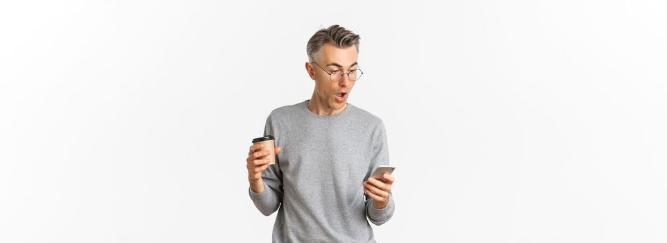 Image of handsome middle-aged man in grey sweater and glasses, drinking coffee and looking amazed at mobile phone screen, reading something online, standing over white background.