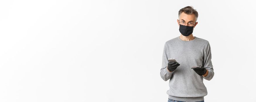 Concept of covid-19, social distancing and lifestyle. Attractive middle-aged guy in medical mask, gloves and glasses, shopping online with smartphone and credit card, standing over white background.