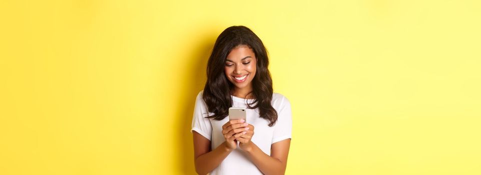 Portrait of good-looking african american girl in white t-shirt, messaging with smartphone, using mobile phone app, standing over yellow background.