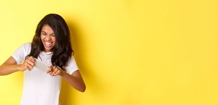 Portrait of cheerful african-american girl, playing mobile game and looking happy, standing over yellow background.