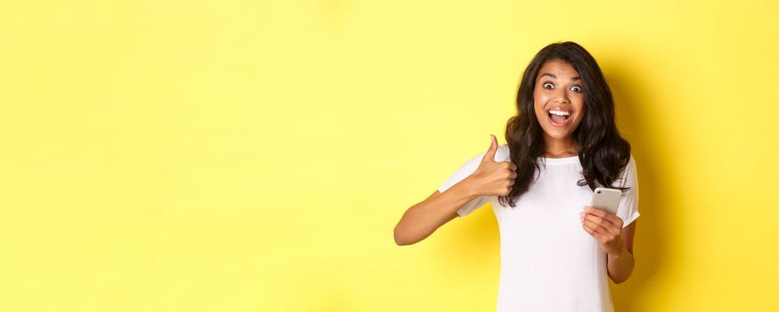 Portrait of amazed african-american girl, showing thumbs-up and using mobile phone, praising cool app, standing over yellow background.