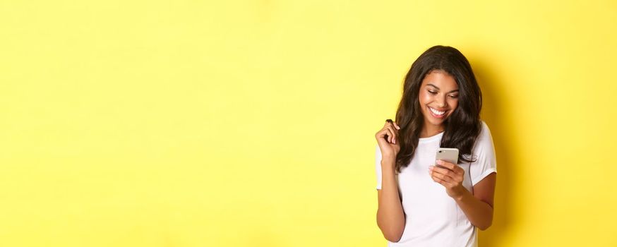 Image of attractive african american girl in white t-shirt, messaging on smartphone, looking at mobile phone and smiling, standing over yellow background.