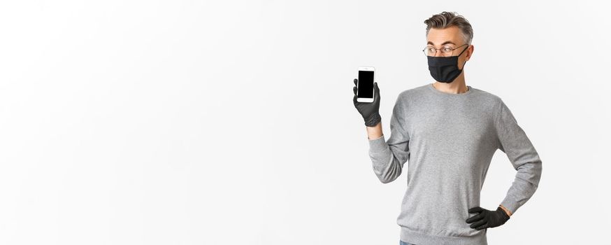 Concept of covid-19, social distancing and lifestyle. Portrait of handsome middle-aged man in medical mask, gloves and glasses, showing mobile phone screen, standing over white background.
