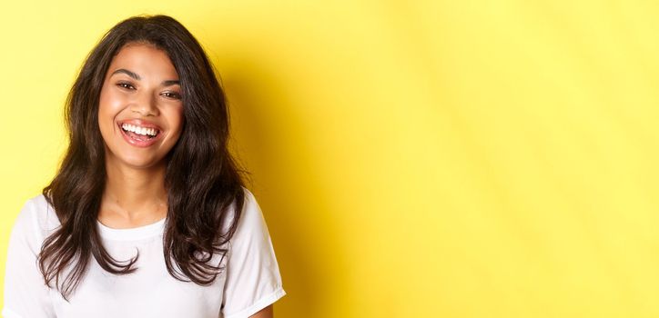 Close-up of joyful and beautiful african-american woman, laughing and looking happy, standing over yellow background.