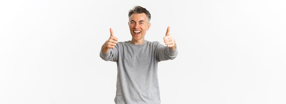 Portrait of cheerful middle-aged man praising good work, standing over white background, showing thumbs-up in approval and smiling glad.