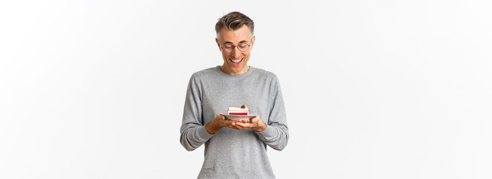 Portrait of handsome middle-aged man, looking happy at delicious cake, standing over white background.