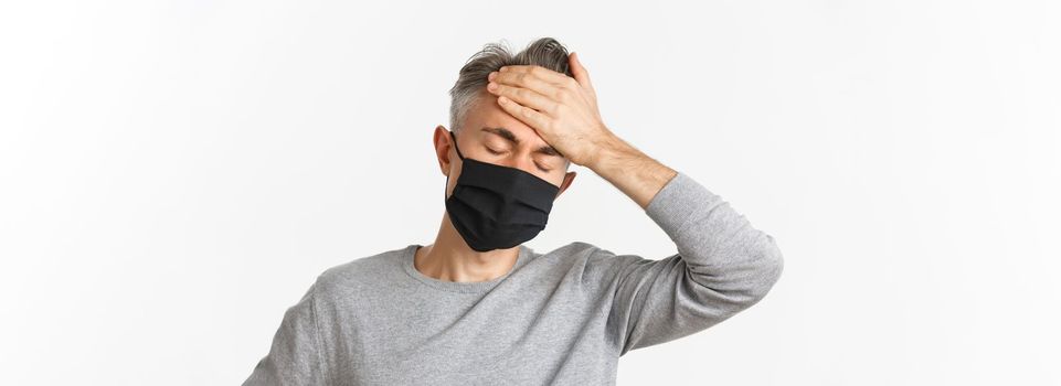 Concept of covid-19, social distancing and quarantine. Close-up of middle-aged man in medical mask, feeling sick or dizzy, touching forehead, suffering headache, have high fever.