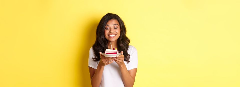 Portrait of beautiful african-american girl celebrating birthday, smiling and looking happy at b-day cake with candle, standing over yellow background.