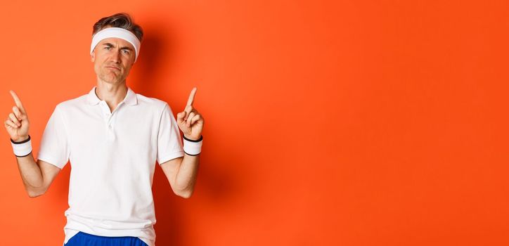 Portrait of skeptical adult sportsman in headband and t-shirt, complaining and pointing fingers up, looking disappointed, standing over orange background.