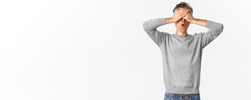 Image of excited middle-aged guy in grey sweater, cover eyes with hands and looking amazed while waiting for surprise, playing hide-n-seek, standing over white background.