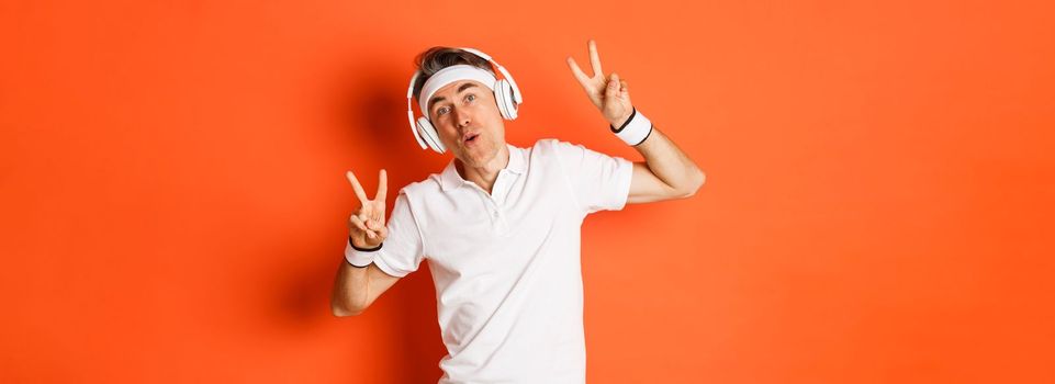 Portrait of carefree middle-aged male athlete, listening music in headphones during fitness training, showing peace signs, standing over orange background.