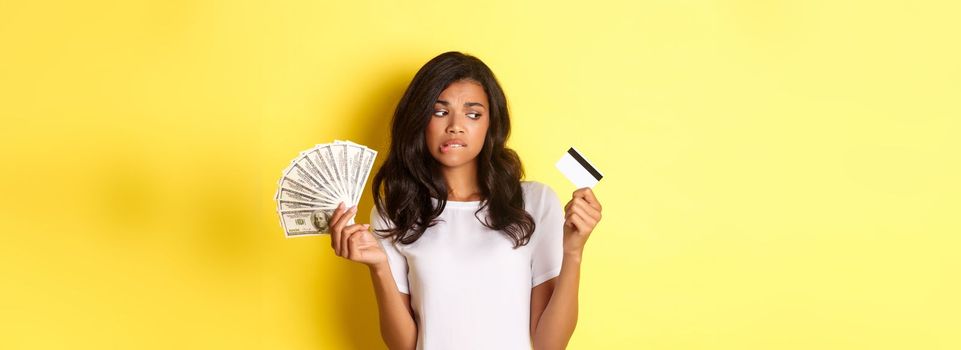 Image of indecisive african-american girl, making choice between money and credit card, standing doubtful over yellow background.