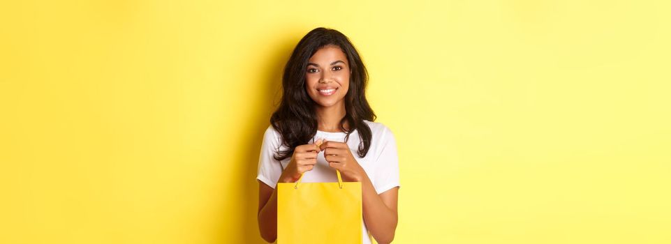 Image of attractive african-american woman, holding shopping baf and smiling, standing over yellow background.
