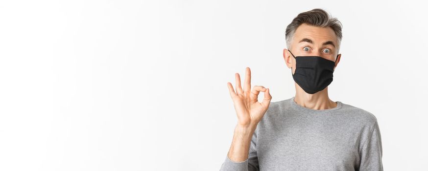 Concept of covid-19, social distancing and quarantine. Close-up of surprised and happy middle-aged man, wearing black medical mask, showing okay sign and look amazed, white background.