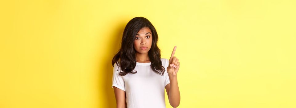 Image of serious african-american girl, scolding someone, raising one finger and making statement, standing over yellow background.