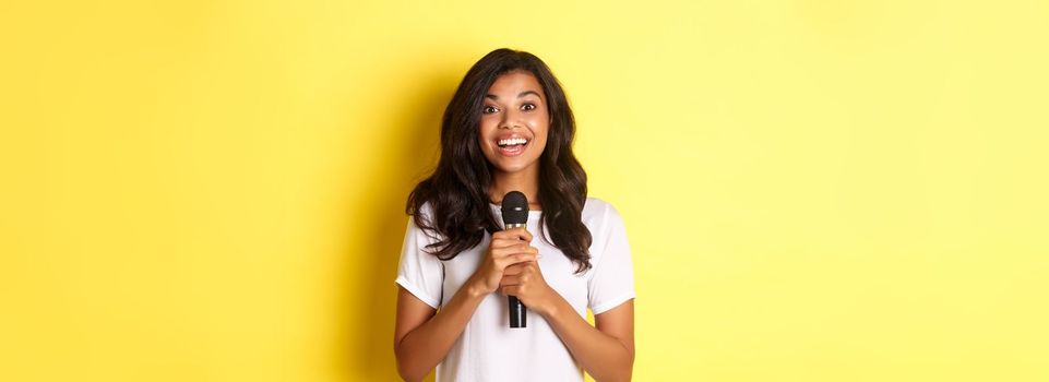 Portrait of happy african-american girl, looking amused while giving speech, holding microphone and smiling at camera, standing over yellow background.