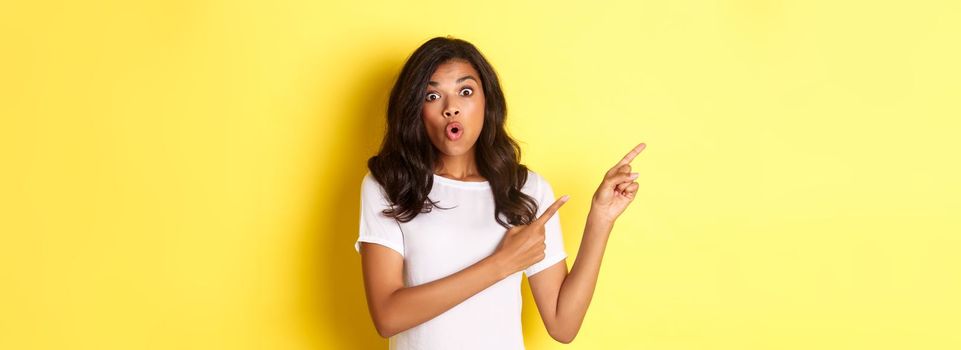 Portrait of surprised african american girl in white t-shirt, looking amazed and pointing fingers at upper left corner, showing your logo or promo banner over yellow background.