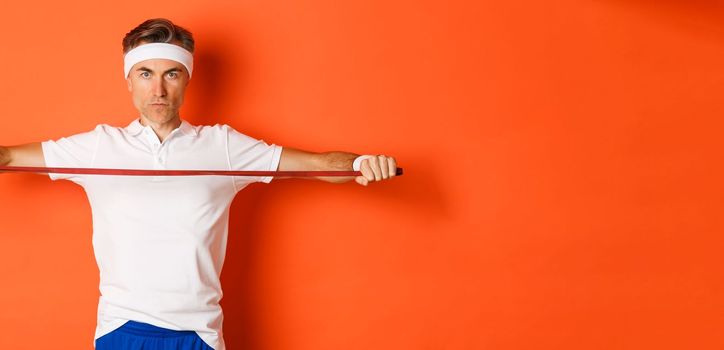 Concept of workout, gym and lifestyle. Serious and confident middle-aged fitness guy, holding stretching rope for exercises, standing over orange background.