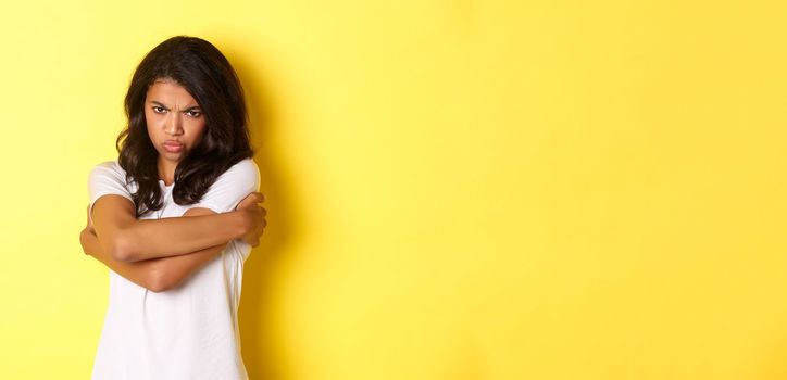 Image of moody cute african-american girl, hugging herself and sulking with offended expression, standing over yellow background.