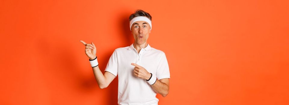 Concept of workout, sports and lifestyle. Portrait of amused middle-aged male athlete, pointing fingers at upper right corner, showing cool promo offer, standing over orange background.