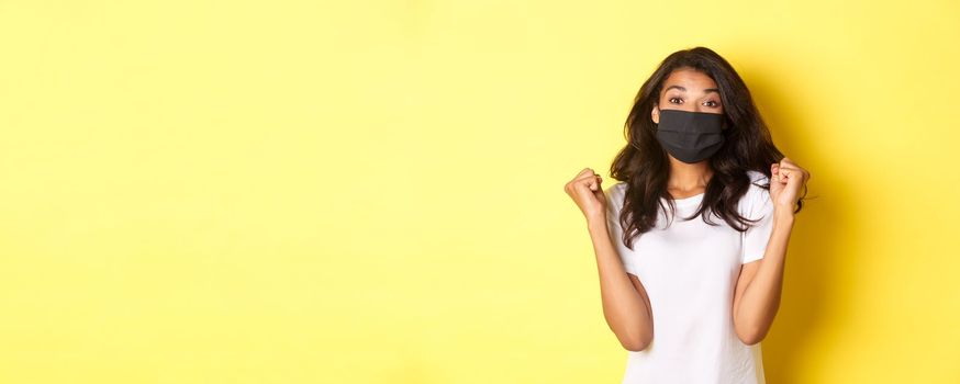 Concept of covid-19, social distancing and lifestyle. Cheerful african-american woman, wearing black face mask, rejoicing with fist pumps and smiling, winning a prize, yellow background.