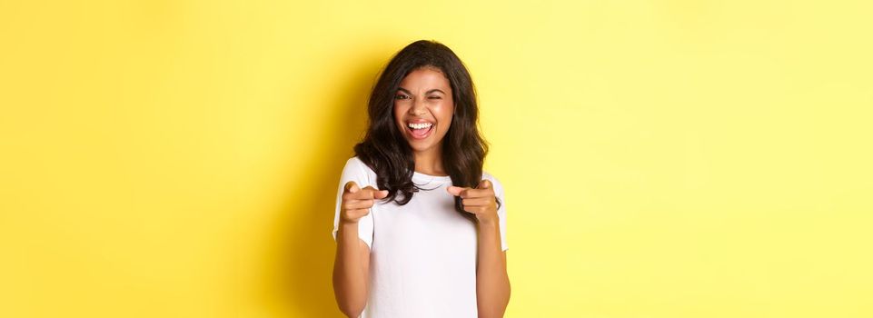 Portrait of cheeky, beautiful african-american woman in white t-shirt, winking and pointing fingers at camera to say congrats, praising you, standing over yellow background.