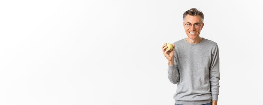 Image of happy handsome middle-aged man, smiling and showing green apple, standing over white background.