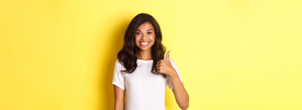 Portrait of happy and cute african-american girl, smiling pleased and showing thumbs-up in approval, like and approve something, standing over yellow background.