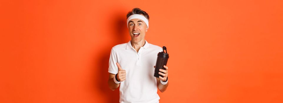 Portrait of healthy and active middle-aged fitness guy, wearing gym uniform, showing thumbs-up, drinking water, standing over orange background.