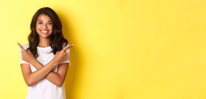 Portrait of happy and confident african-american girl showing two choices, pointing fingers sideways at right and left copy space, smiling pleased, standing over yellow background.