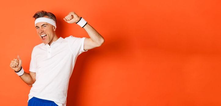 Portrait of cheerful middle-aged male athlete, become champion, raising hands up and shouting for joy, doing sports over orange background.