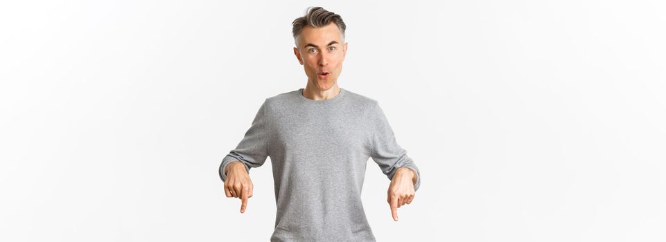 Surprised handsome middle-aged guy in grey sweater asking question about product, pointing fingers down and looking amazed, standing over white background.