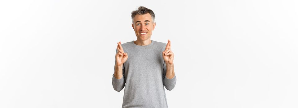 Image of excited and hopeful middle-aged man making a wish, waiting for results, cross fingers for good luck and smiling, standing over white background.