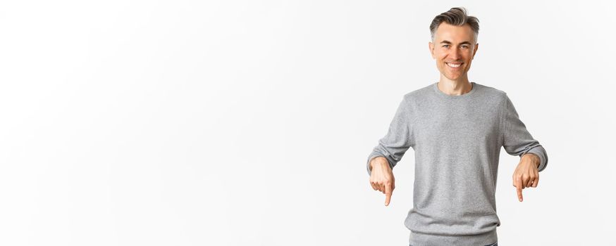 Handsome and confident middle-aged man in grey sweater, pointing fingers down and smiling, showing good deal promo, standing over white background.