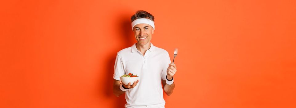 Concept of sport, fitness and lifestyle. Image of handsome, healthy and active male athlete, eating salad and smiling, standing over orange background.