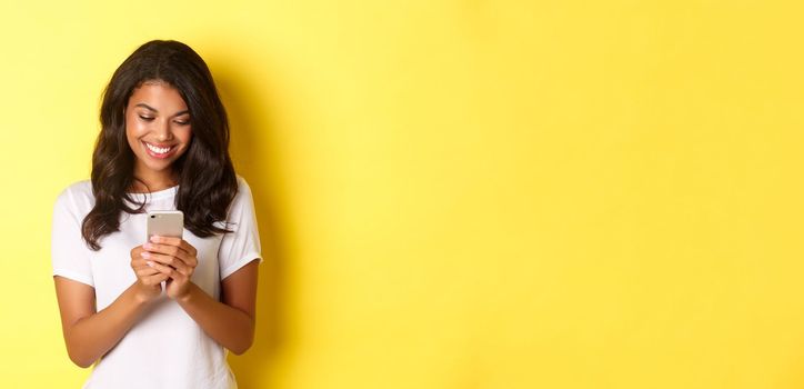 Portrait of good-looking african american girl in white t-shirt, messaging with smartphone, using mobile phone app, standing over yellow background.