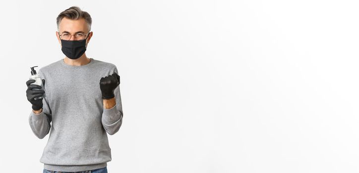 Concept of coronavirus, lifestyle and quarantine. Image of cheerful caucasian man in medical mask and gloves, showing hand sanitizer and making fist pump to rejoice, standing over white background.