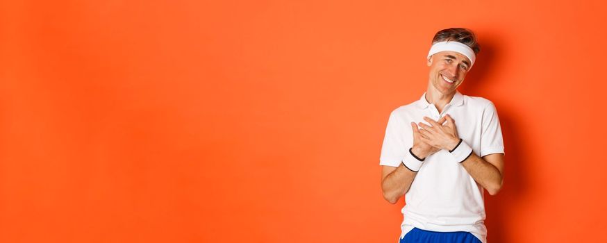 Concept of workout, sports and lifestyle. Portrait of touched and grateful adult man in sportswear, thanking and looking pleased, holding hands on heart and smiling, orange background.
